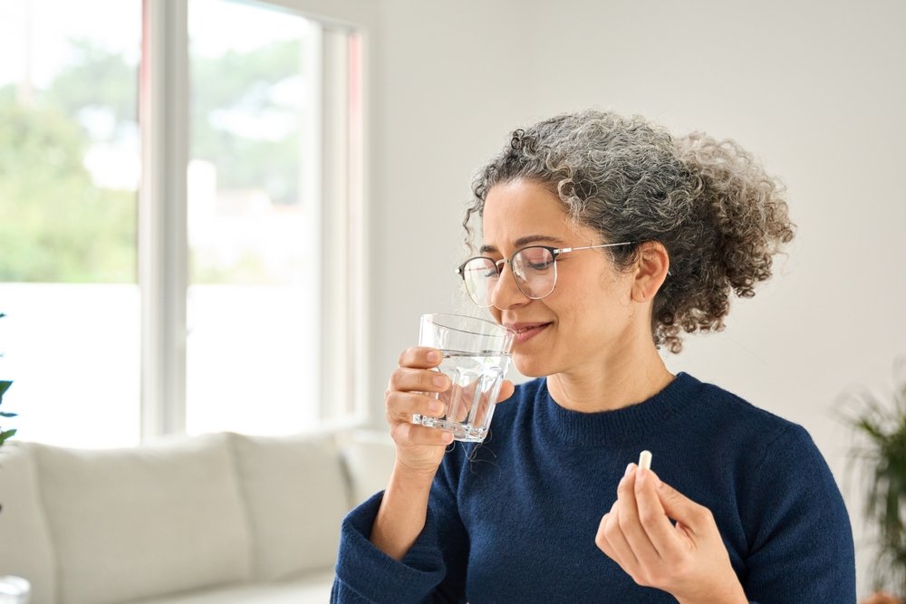 Mujer sana feliz de la píldora de calcio que habla de la mediana edad que sostiene el vaso del agua que se coloca en casa.